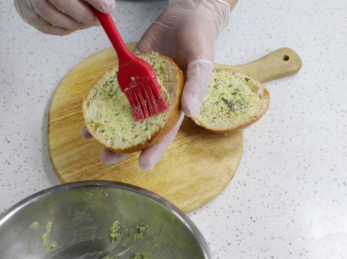 Une main gantée utilisant un pinceau en silicone rouge pour étaler uniformément le beurre à lail sur la moitié dun pain à burger coupé sur une planche en bois.