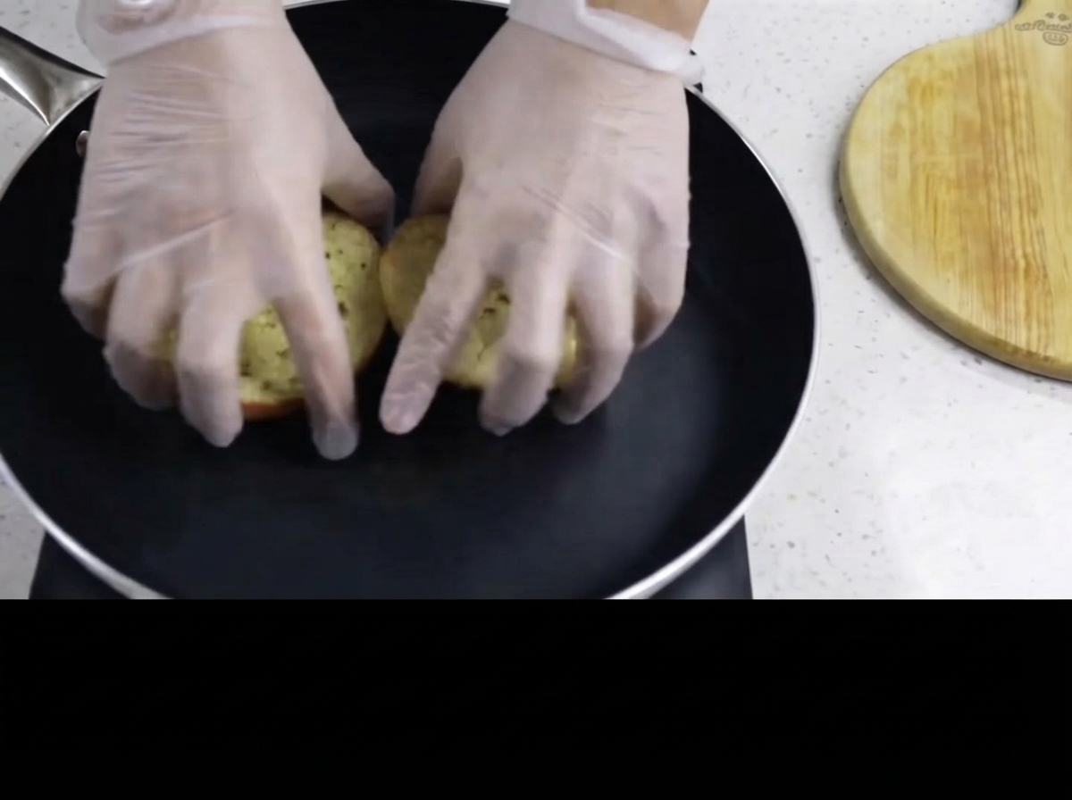 Des mains gantées plaçant le côté beurré des pains à burger face vers le bas dans une poêle noire chaude.