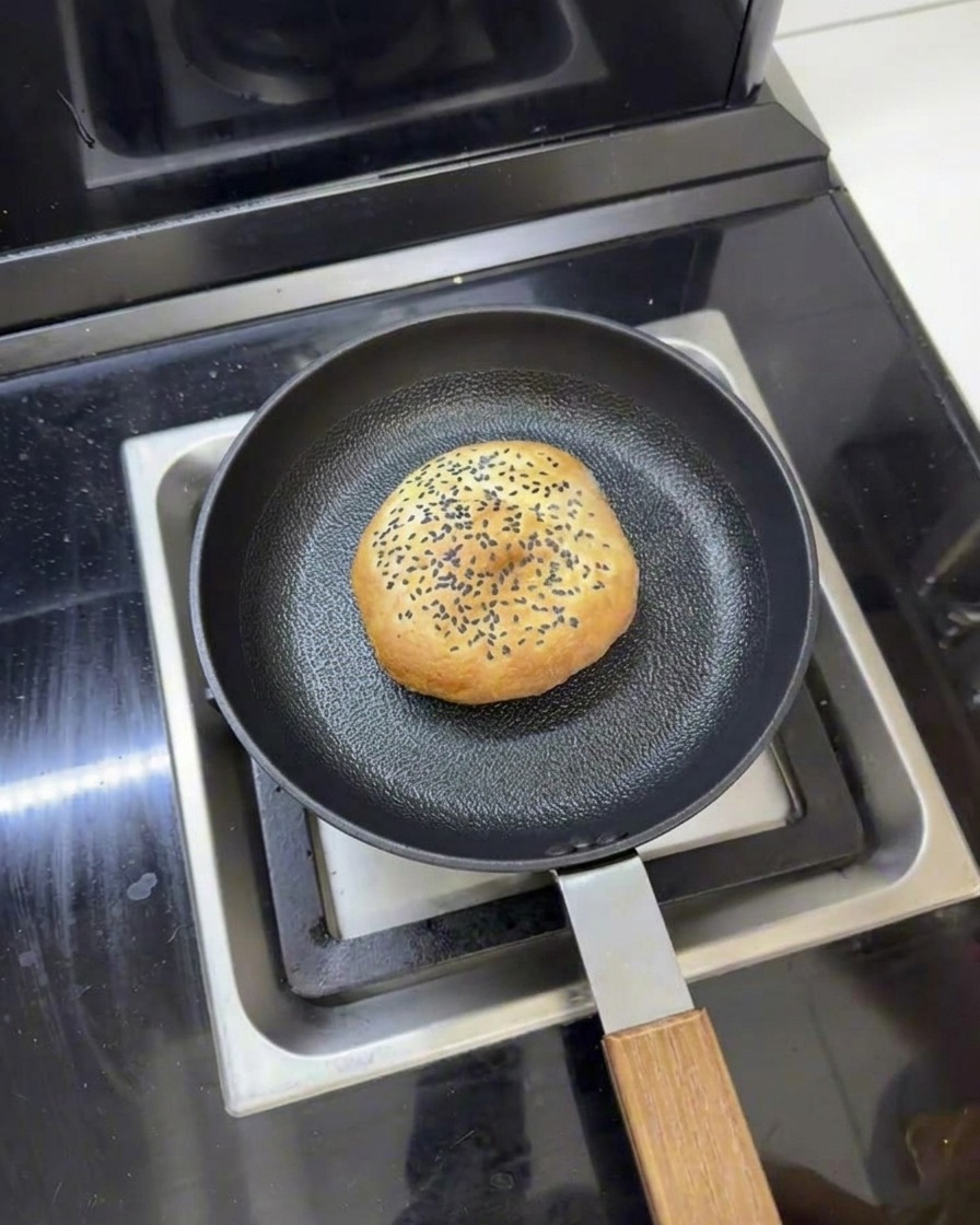 Un pain à burger au beurre dans une poêle avec de leau autour, prêt à être réchauffé à la vapeur avec un couvercle.