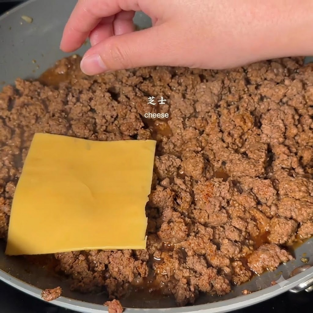 Une tranche de fromage carrée jaune placée sur le dessus du bœuf haché cuit dans une poêle par une main.
