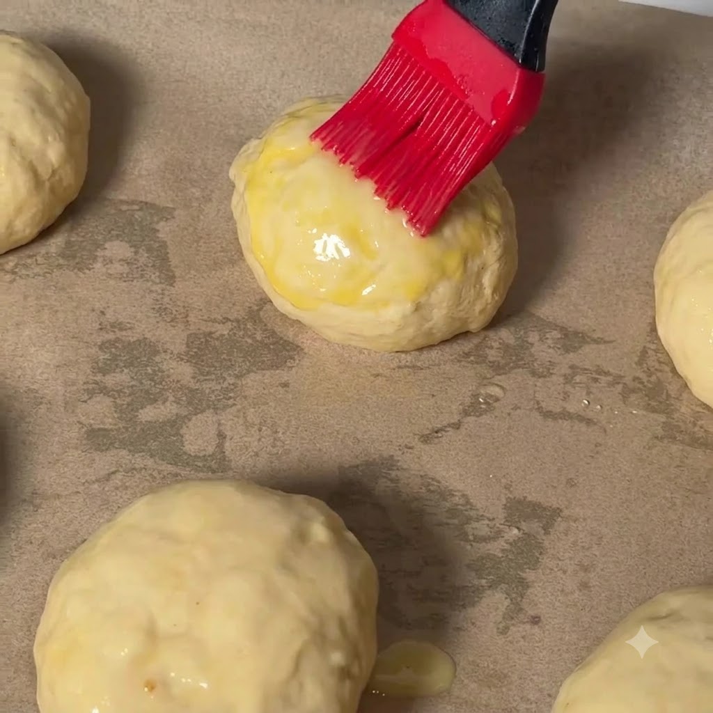 Un pinceau à pâtisserie rouge appliquant une dorure à lœuf sur le dessus dune boule de pâte à cheeseburger sur du papier sulfurisé.