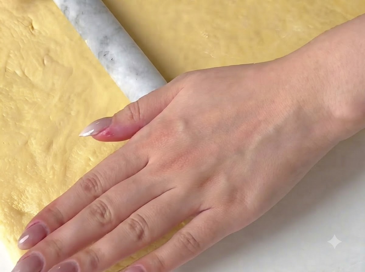 Une main utilisant un rouleau à pâtisserie en marbre pour aplatir une feuille de pâte jaune sur une surface.