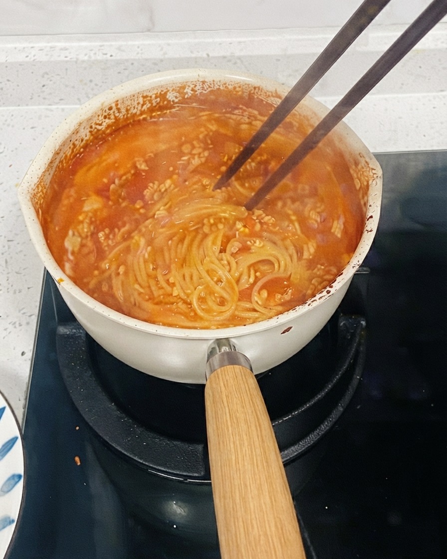 Une casserole de pâtes à base de tomate mijotant sur une cuisinière avec des baguettes remuant les nouilles pour épaissir la sauce.