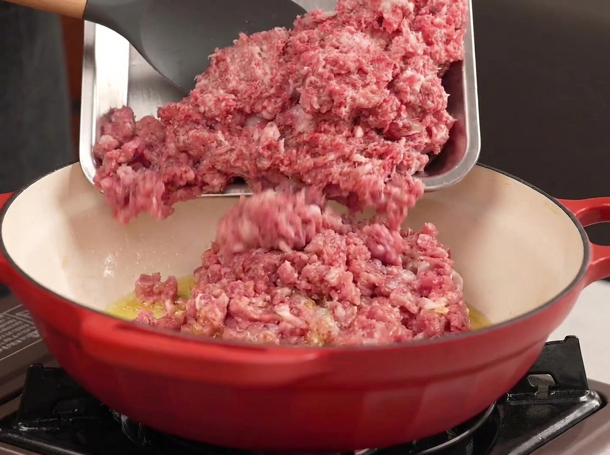 Du bœuf et du porc hachés versés dun plateau en métal dans une casserole rouge avec de lhuile chaude.