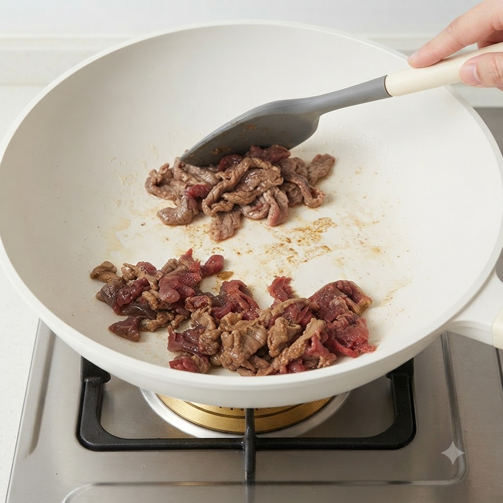 Tranches de bœuf marinées en train dêtre sautées dans une poêle blanche à laide dune spatule en silicone grise.