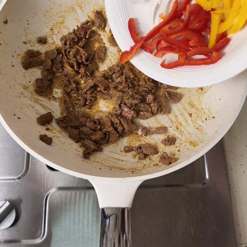 Poivrons rouges et jaunes tranchés versés dune assiette blanche dans une poêle contenant des tranches de bœuf cuites.