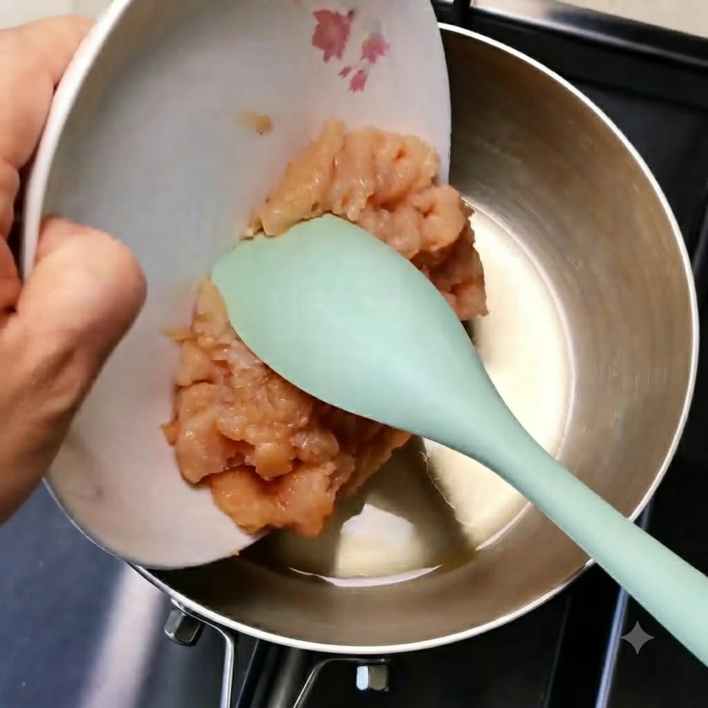 De la viande de poulet hachée glissant dune assiette blanche vers une poêle en métal à laide dune spatule verte.