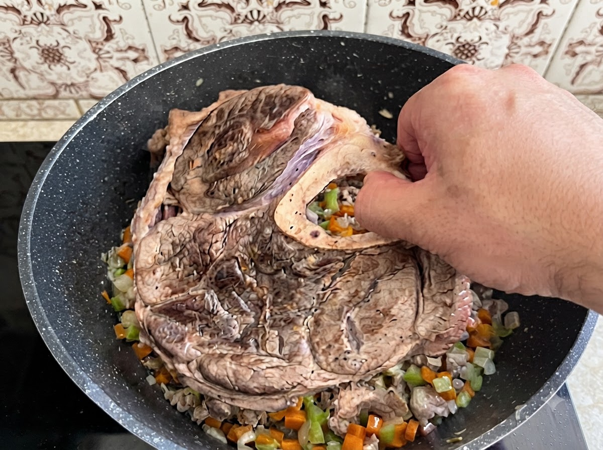 Une main plaçant un grand morceau de plat de côtes de bœuf saisi et son os dans une cocotte remplie de légumes cuits en dés.