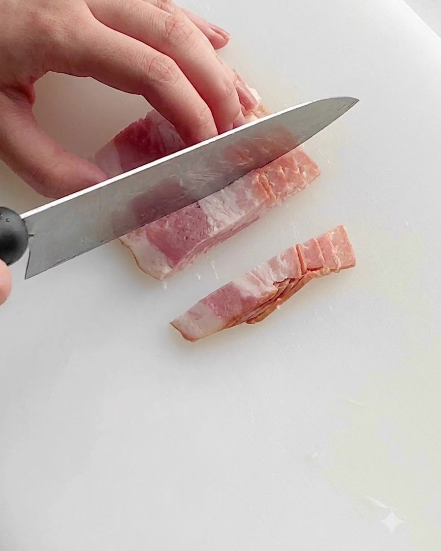 Un couteau de cuisine coupant des tranches de bacon cru en petits morceaux réguliers sur une planche à découper blanche en plastique.