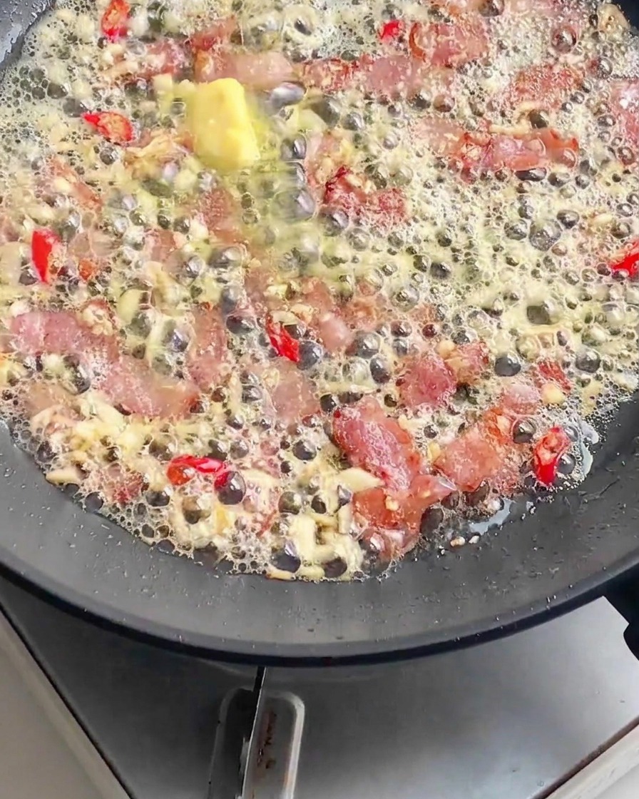 Des nouilles de pâtes cuites ajoutées dans une poêle remplie de bacon frémissant, de beurre fondu, dail émincé et de piments rouges tranchés.