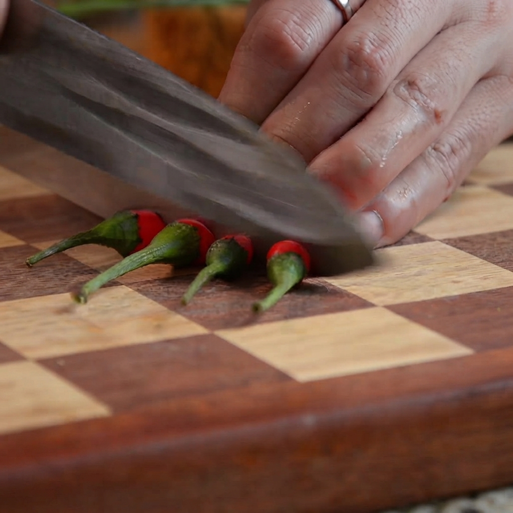 Un couteau de cuisine coupant des piments rouges en petits morceaux sur une planche à découper en bois.