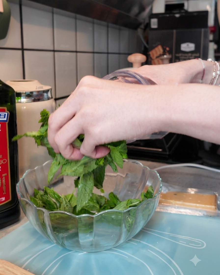 Feuilles de basilic frais ajoutées dans un bol de mixeur transparent avec de lail et des pignons de pin.