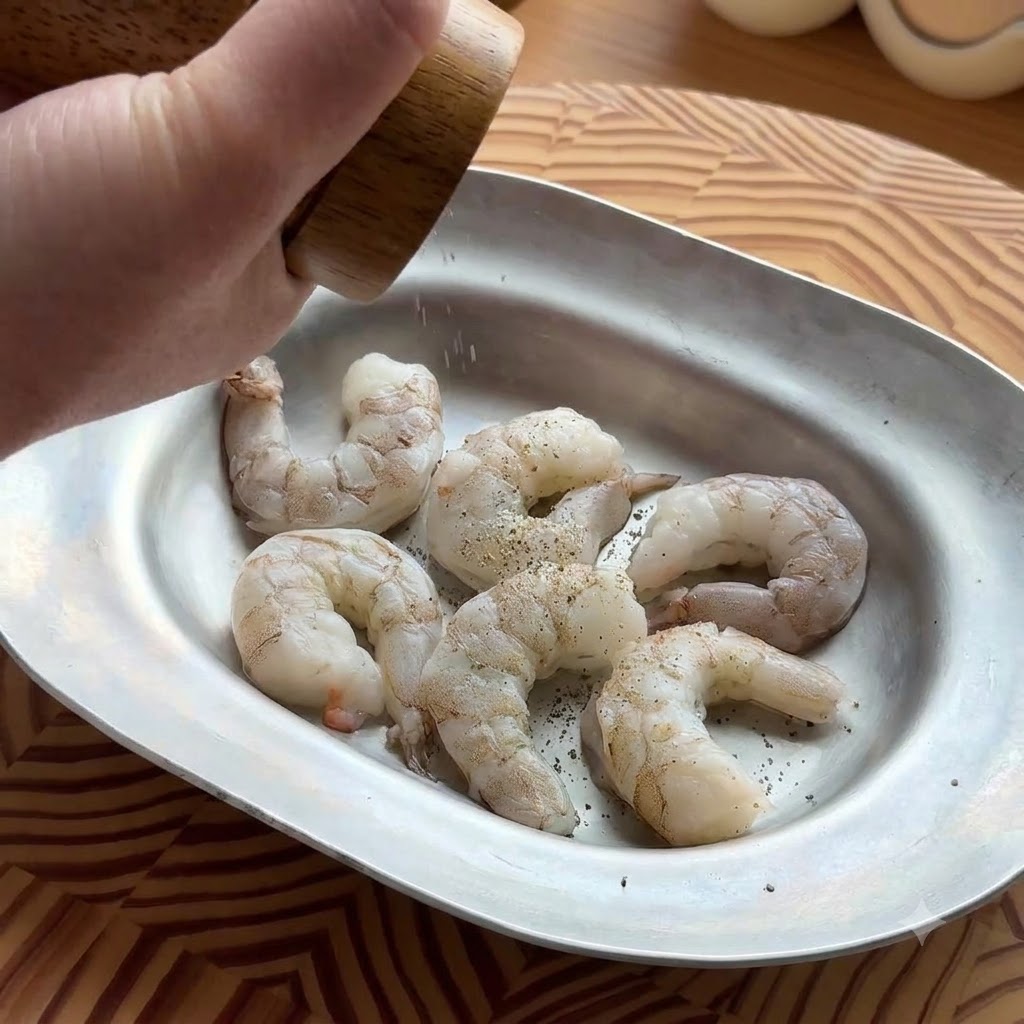 Une main utilisant un moulin à poivre en bois pour assaisonner des crevettes crues et décortiquées dans un plat métallique ovale.