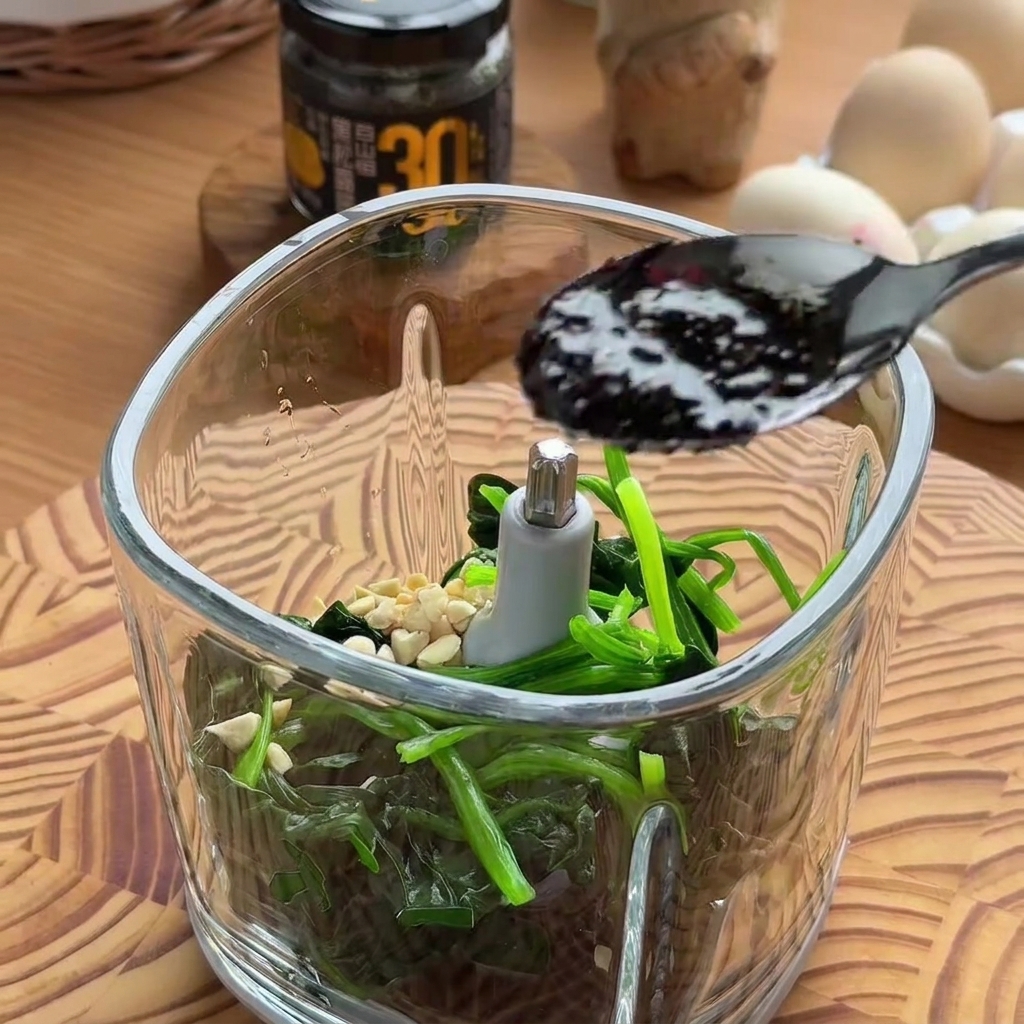 Une cuillère noire versant de la sauce à la truffe noire sombre et riche dans le bol dun robot culinaire rempli dépinards et de fruits à coque.