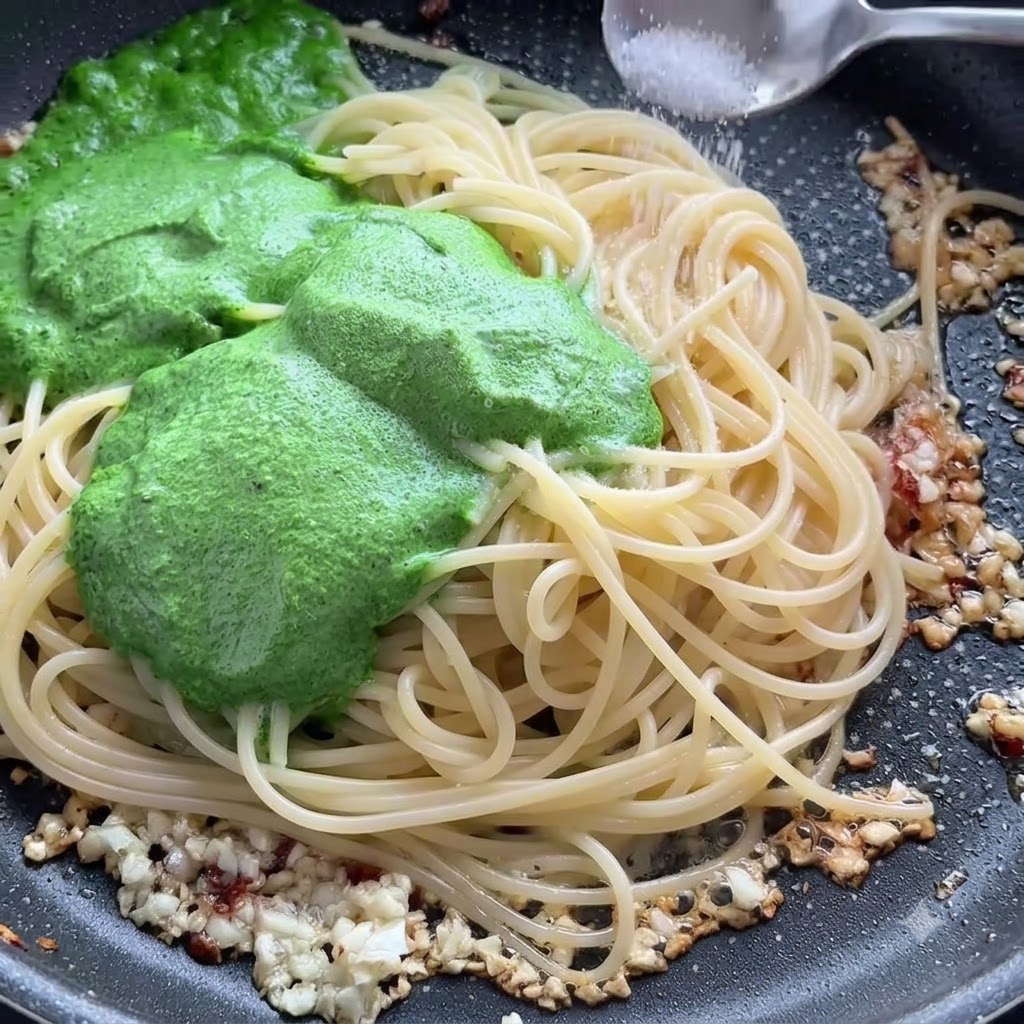 Une pincée de sel saupoudrée sur des spaghettis et de la sauce pesto verte dans une poêle.