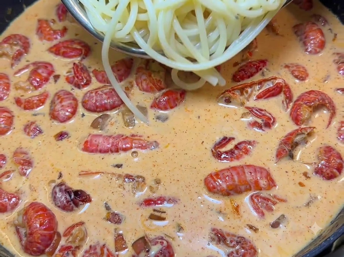 Des pinces soulevant des nouilles spaghetti bouillies dans une poêle grésillante remplie de sauce rose crémeuse et de queues décrevisses rouges.