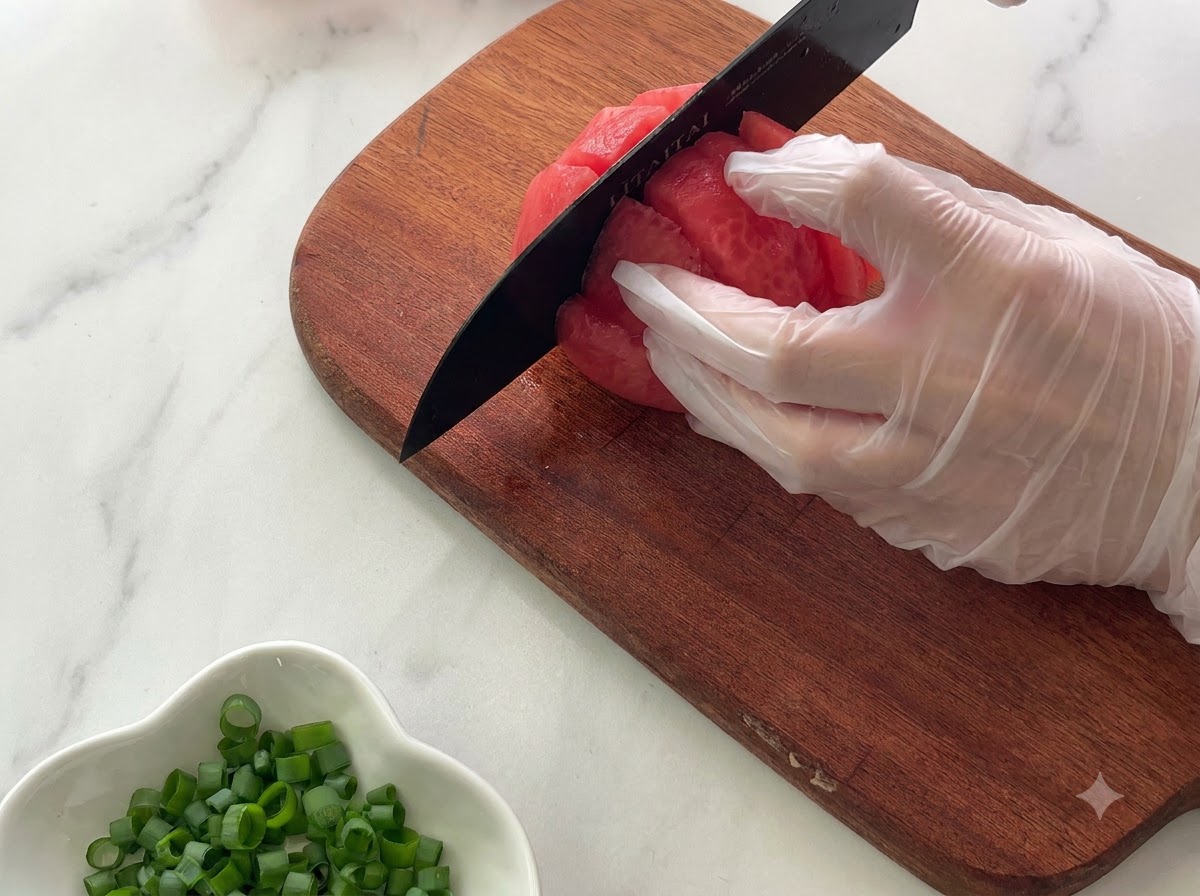 Une main gantée tenant une tomate sur une planche à découper en bois tout en la coupant en morceaux avec un couteau noir.