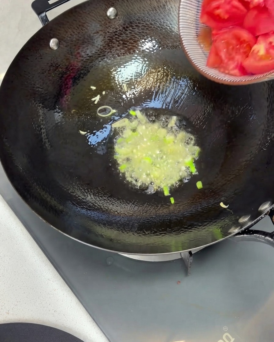 Morceaux de tomates versés dun bol dans un wok avec des oignons verts sautés et de lhuile.