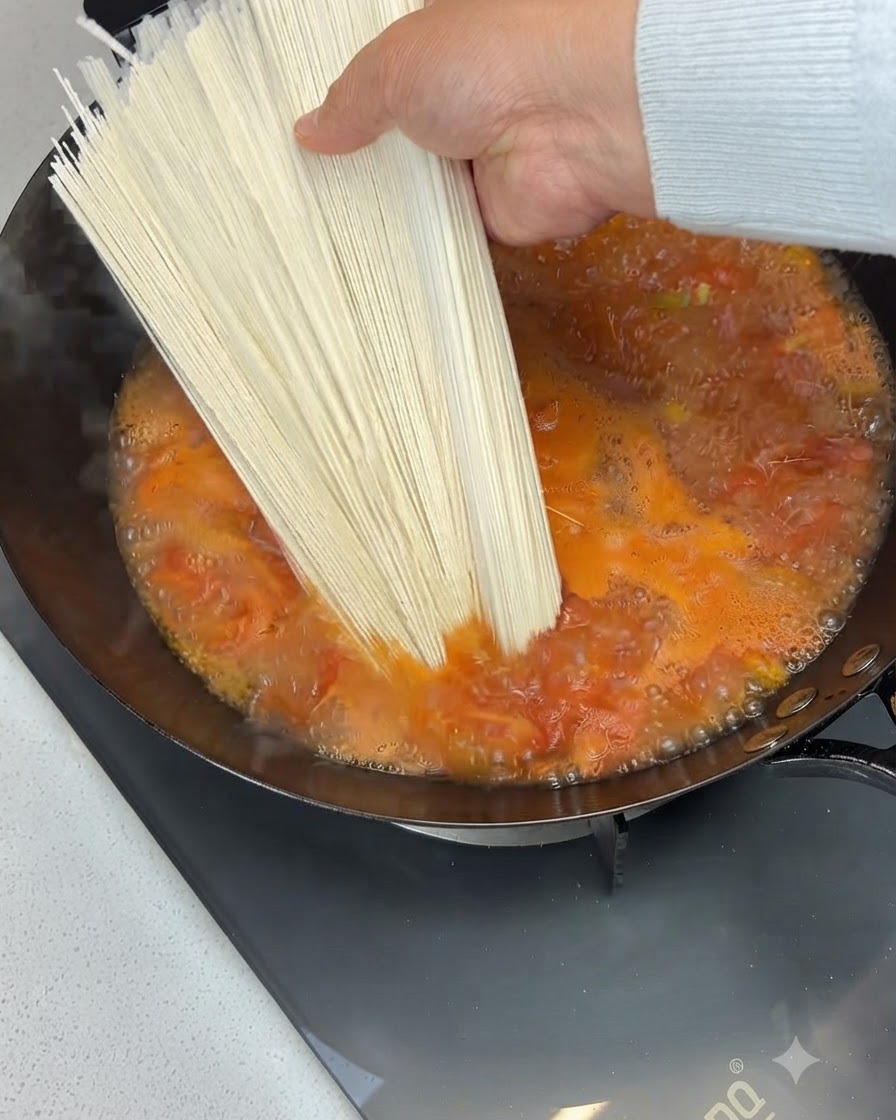 Une botte de nouilles sèches tenue au-dessus et ajoutée dans une soupe à la tomate bouillonnante dans un wok.