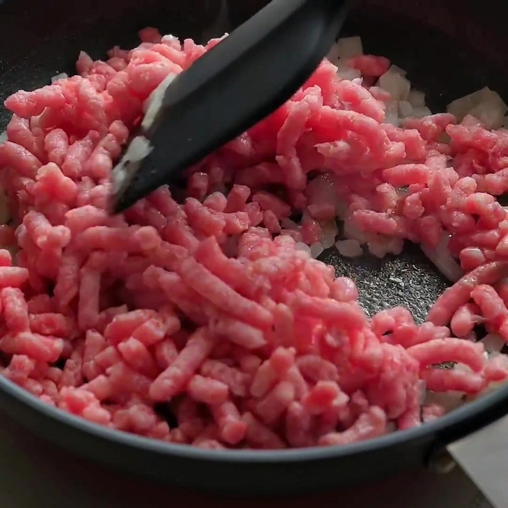 Du bœuf haché rose cru émietté et sauté avec des oignons dans une poêle noire.