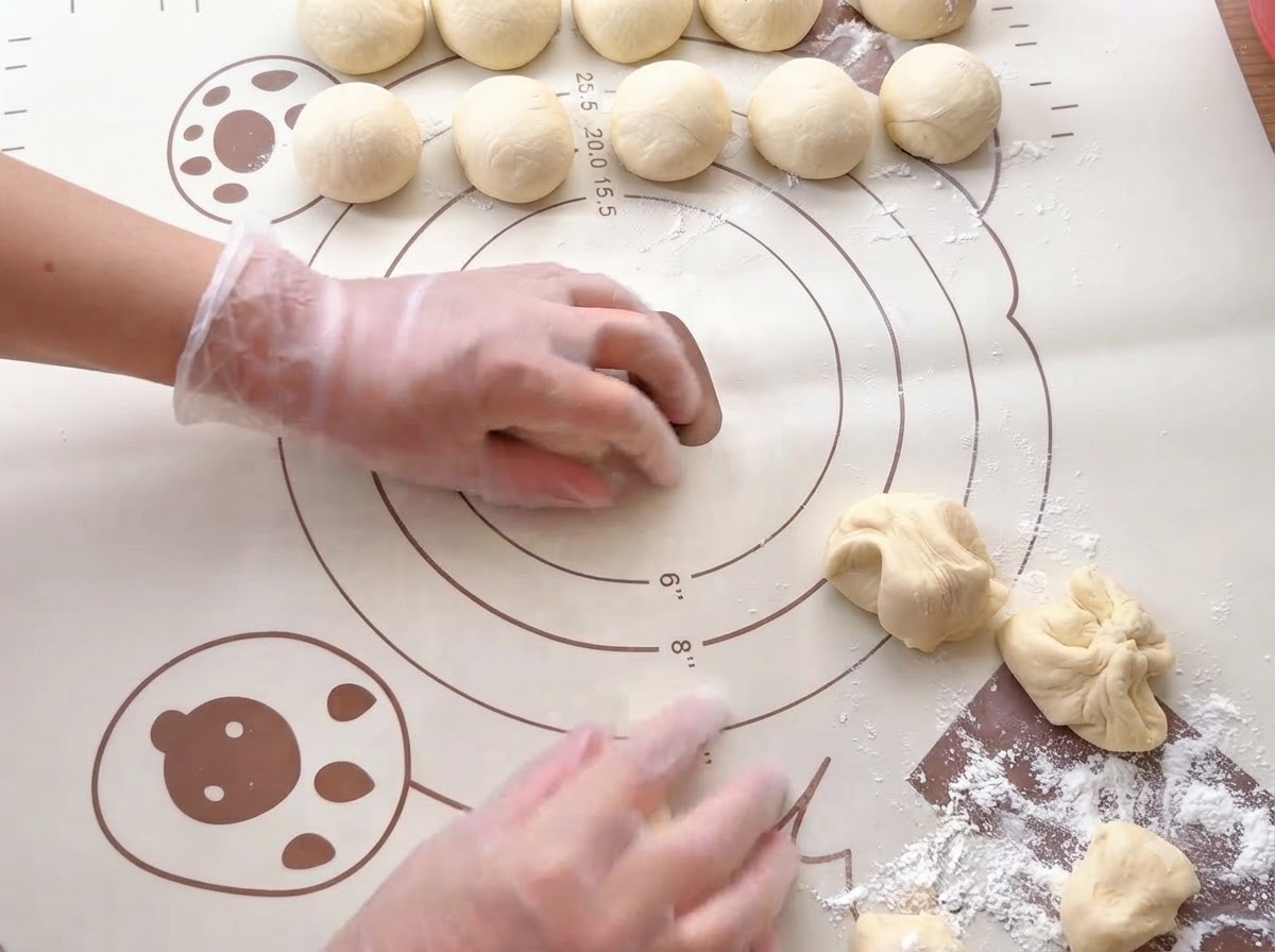 Des mains roulant de petites portions de pate en boules lisses sur un tapis de cuisson en silicone imprime.