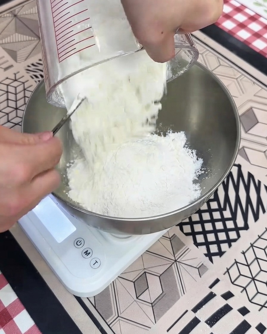 De la farine riche en protéines et sèche versée dune tasse à mesurer dans un bol en métal posé sur une balance numérique blanche.