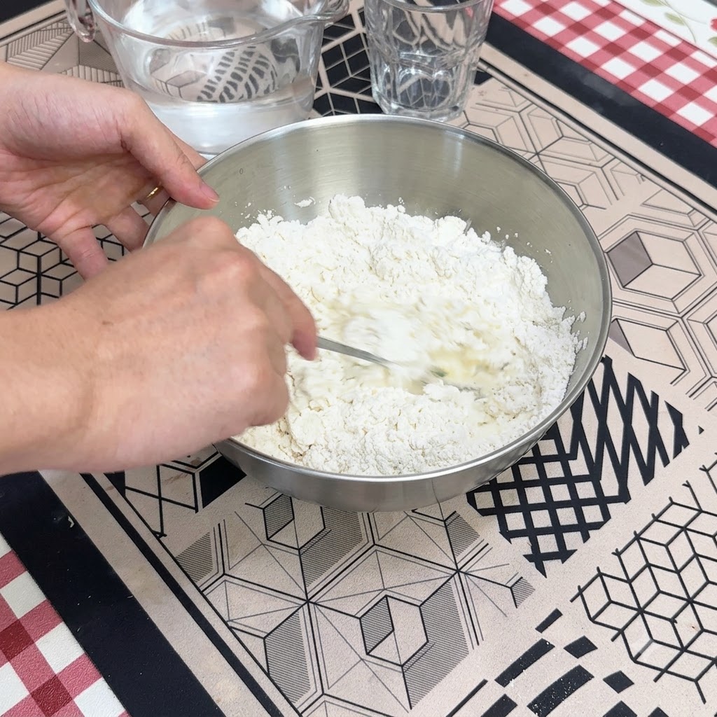 Une main utilisant une cuillère pour mélanger de leau avec de la farine sèche à lintérieur dun bol mélangeur en métal.