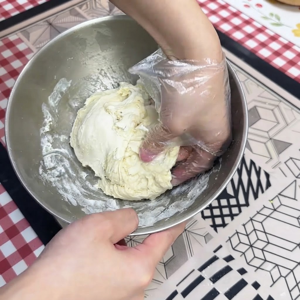 Une main gantée pétrissant de la pâte à pizza humide à lintérieur dun bol mélangeur en acier inoxydable.