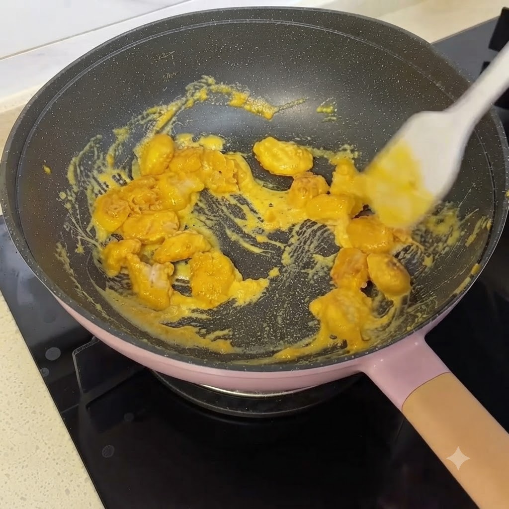 Des morceaux de poulet pop-corn en train dêtre mélangés et enrobés de sauce jaune aux jaunes dœufs salés dans une poêle à laide dune spatule en silicone.