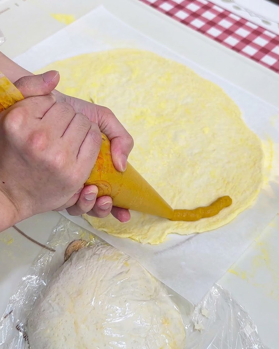 Une paire de mains utilisant une poche à douille pour pocher de la purée de patate douce jaune autour du bord dune feuille de pâte à pizza plate sur du papier sulfurisé.