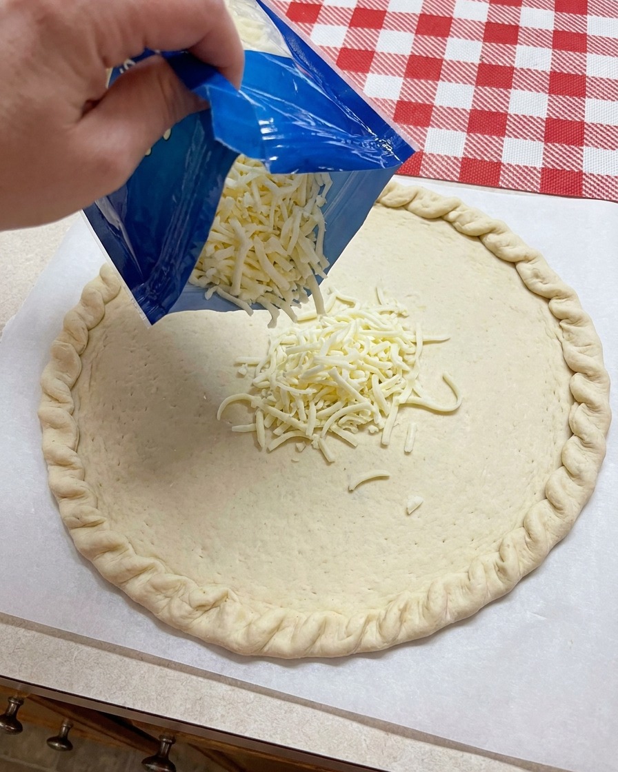 Du fromage mozzarella râpé versé dun sachet bleu sur le centre dune base de pâte à pizza façonnée.