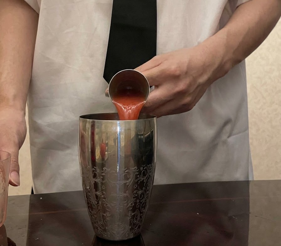 Du jus de tomate rouge frais verse depuis un jigger en metal dans un shaker a cocktail argente et etnampé.