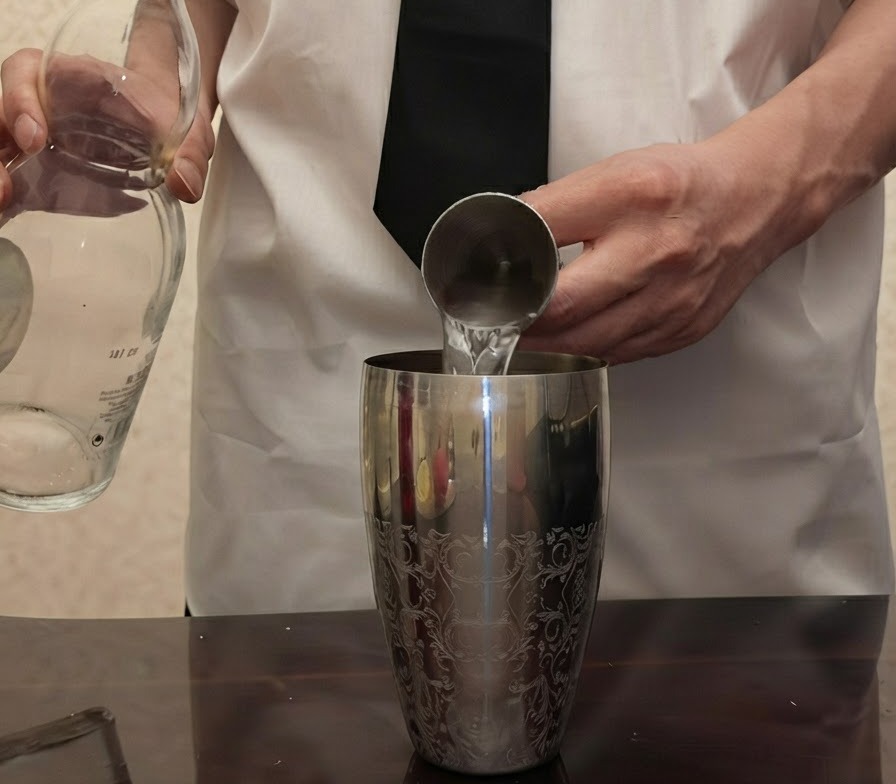 De la tequila transparente versee dune bouteille en verre dans un jigger en metal tenu au-dessus dun shaker a cocktail en argent.