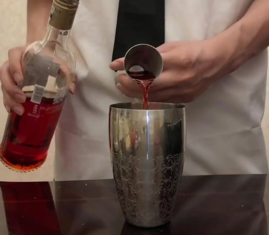 Du Campari rouge vif verse de sa bouteille dans un jigger en metal au-dessus dun shaker a cocktail en argent.
