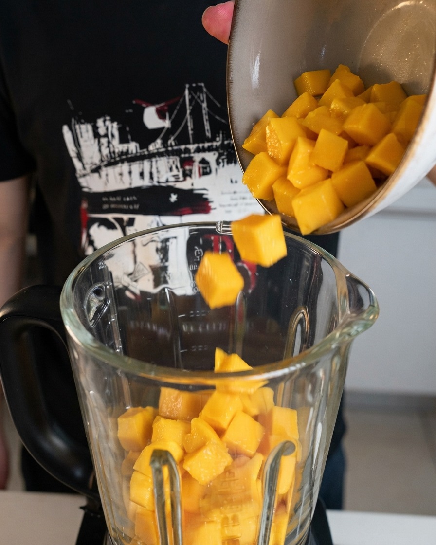Des cubes de mangue jaune fraîchement coupés versés dun bol dans le bol dun blender transparent.