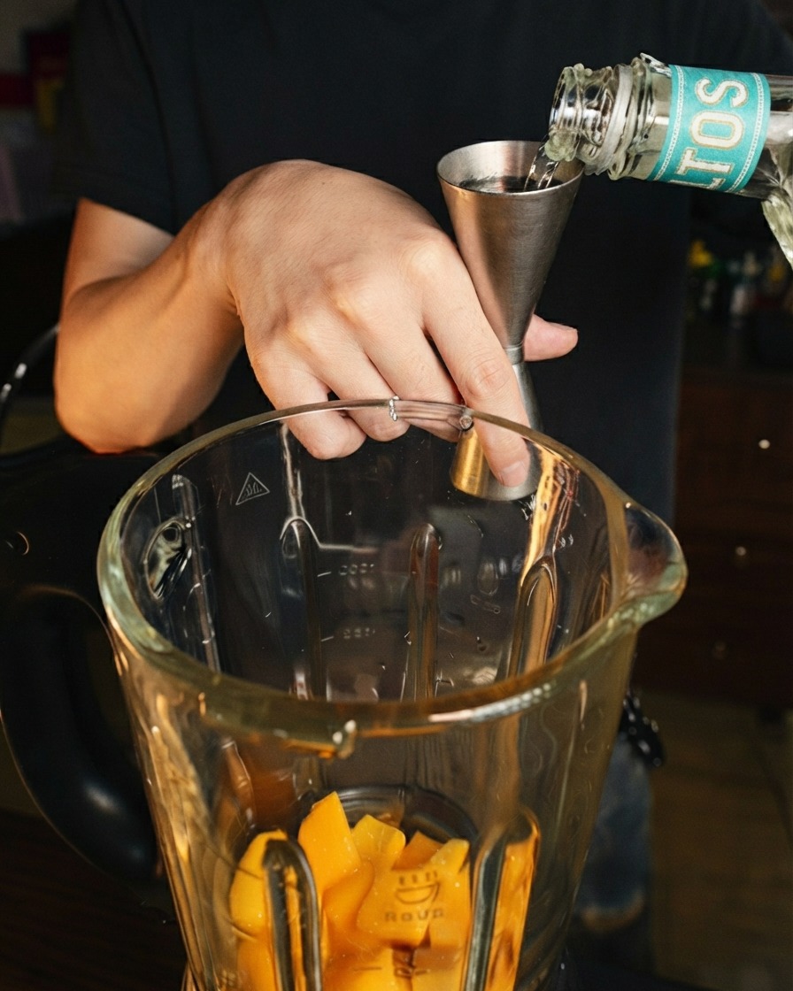 De lalcool clair versé dun jigger en acier inoxydable dans le bol du blender par-dessus les morceaux de mangue.