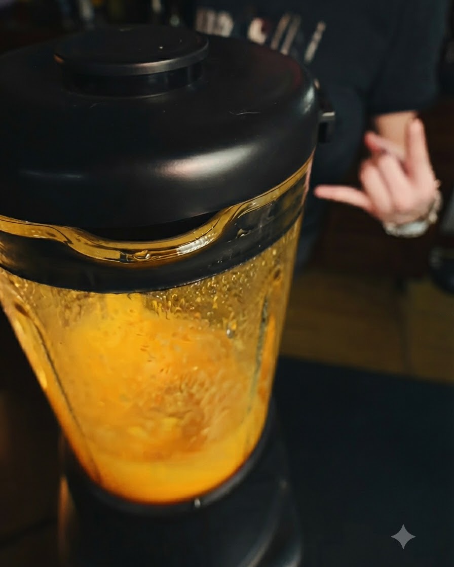 Un blender en marche contenant une préparation de mangue dun orange vif, avec une main faisant un geste en arrière-plan.