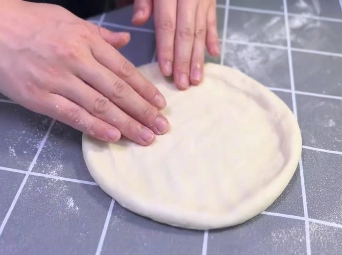 Mains étirant et façonnant doucement une boule de pâte à pizza huilée sur une surface quadrillée grise.
