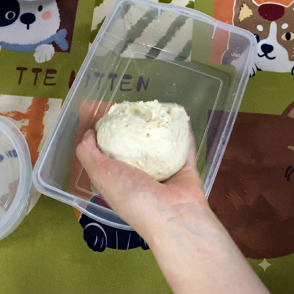 Une main plaçant une boule de pâte lisse et ronde dans un contenant de bac à légumes en plastique rectangulaire.