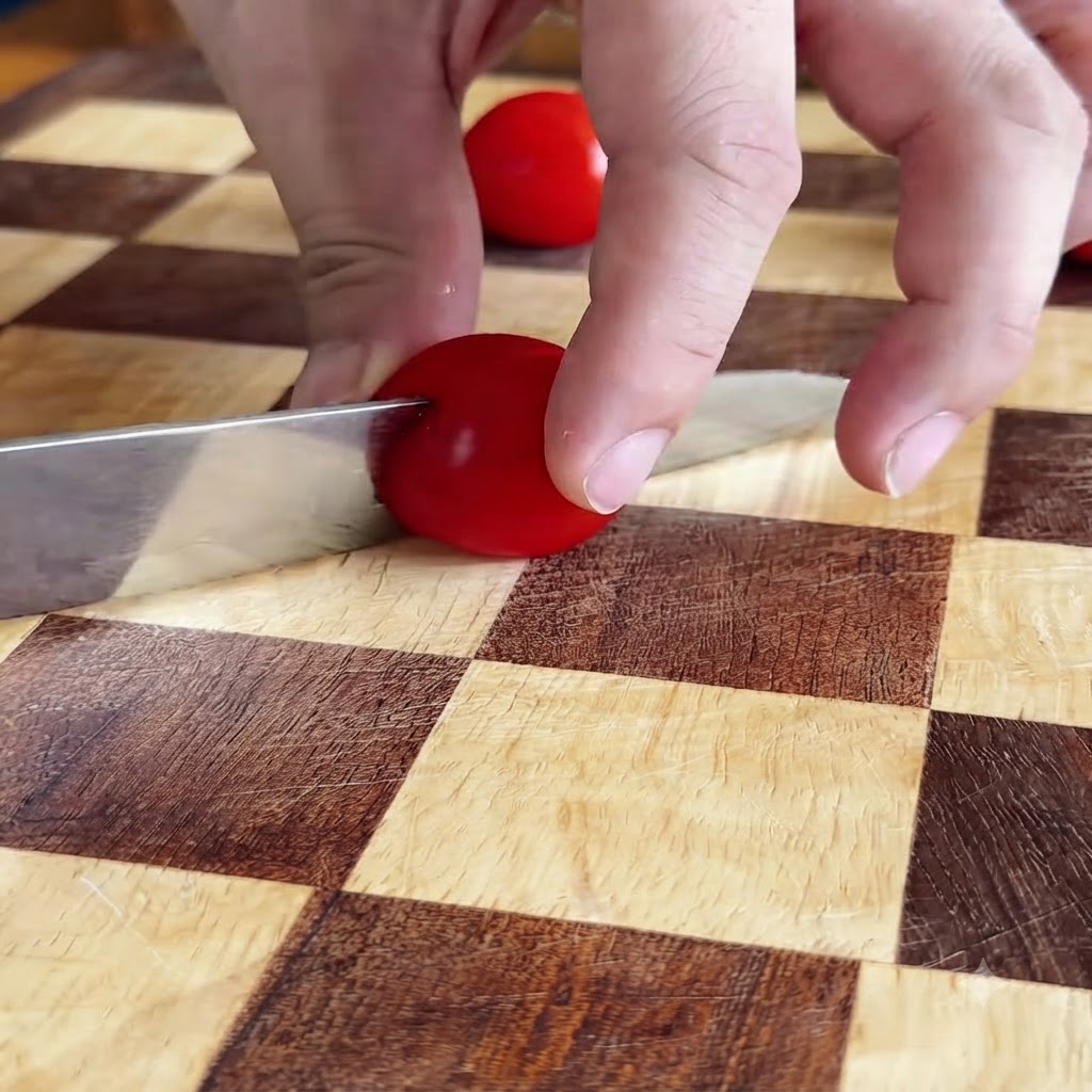 Mains coupant une tomate cerise rouge en deux avec un couteau sur une planche à découper à damier.