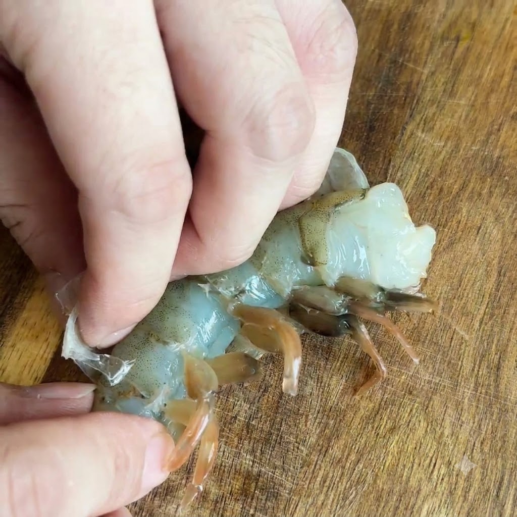 Doigts retirant la carapace translucide dune crevette crue sur une surface en bois.
