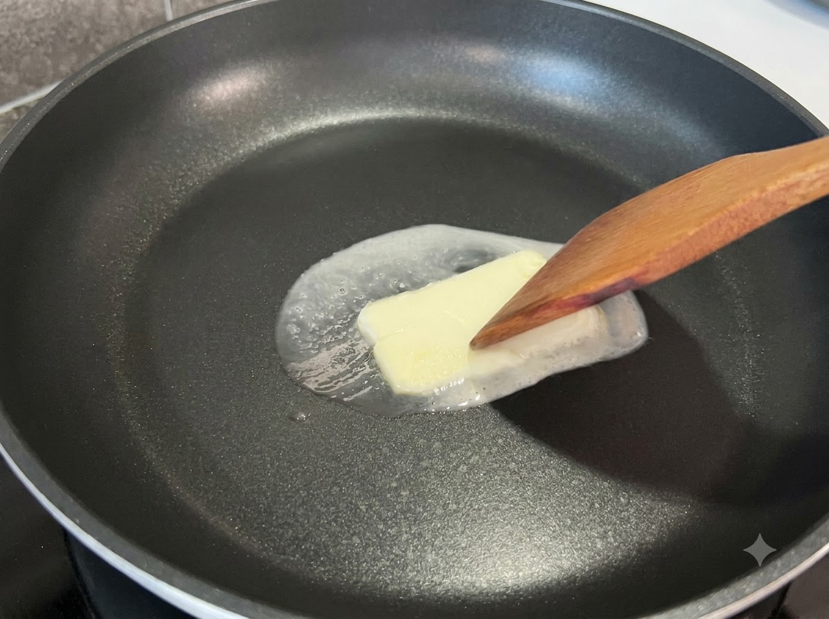 Une spatule en bois appuyant légèrement sur un carré de beurre fondant dans une poêle noire.