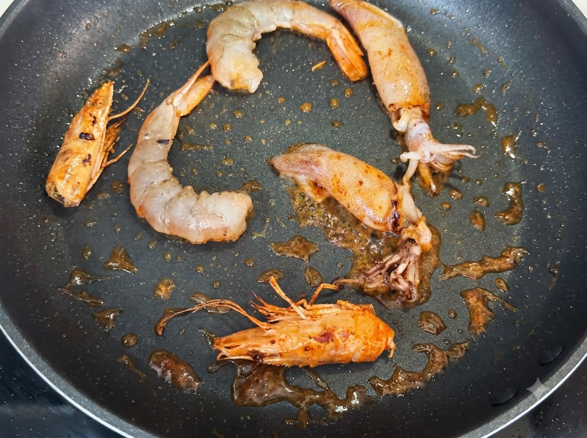 Crevettes, têtes de crevettes entières et morceaux de calmar en train de frire dans une poêle, assaisonnés de petits grains noirs de poivre.