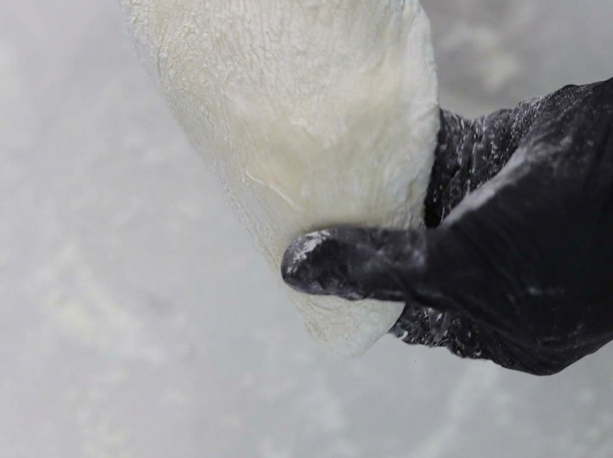 Des mains portant des gants noirs qui étirent et façonnent la pâte à pizza sur une surface farinée.
