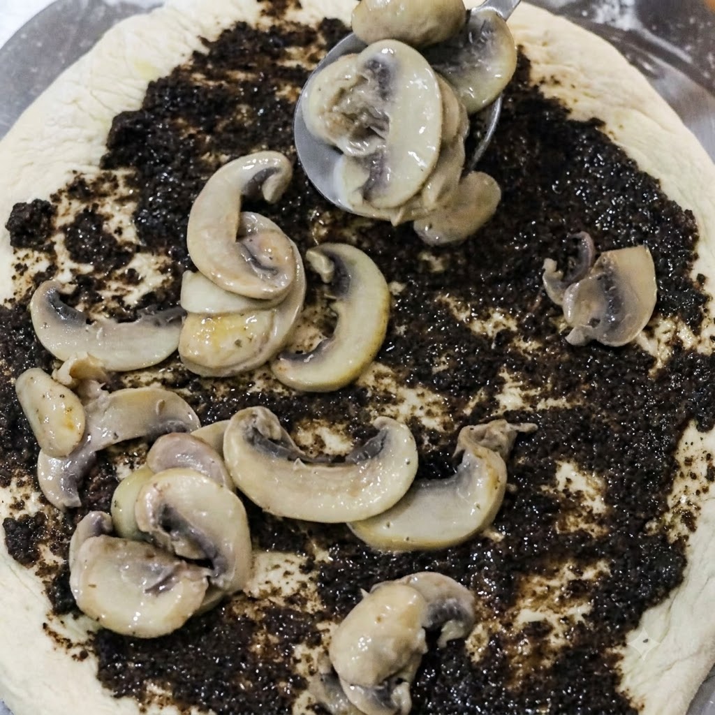 Une cuillère déposant des tranches de champignons cuits sur une base de pizza recouverte de crème de truffe noire.
