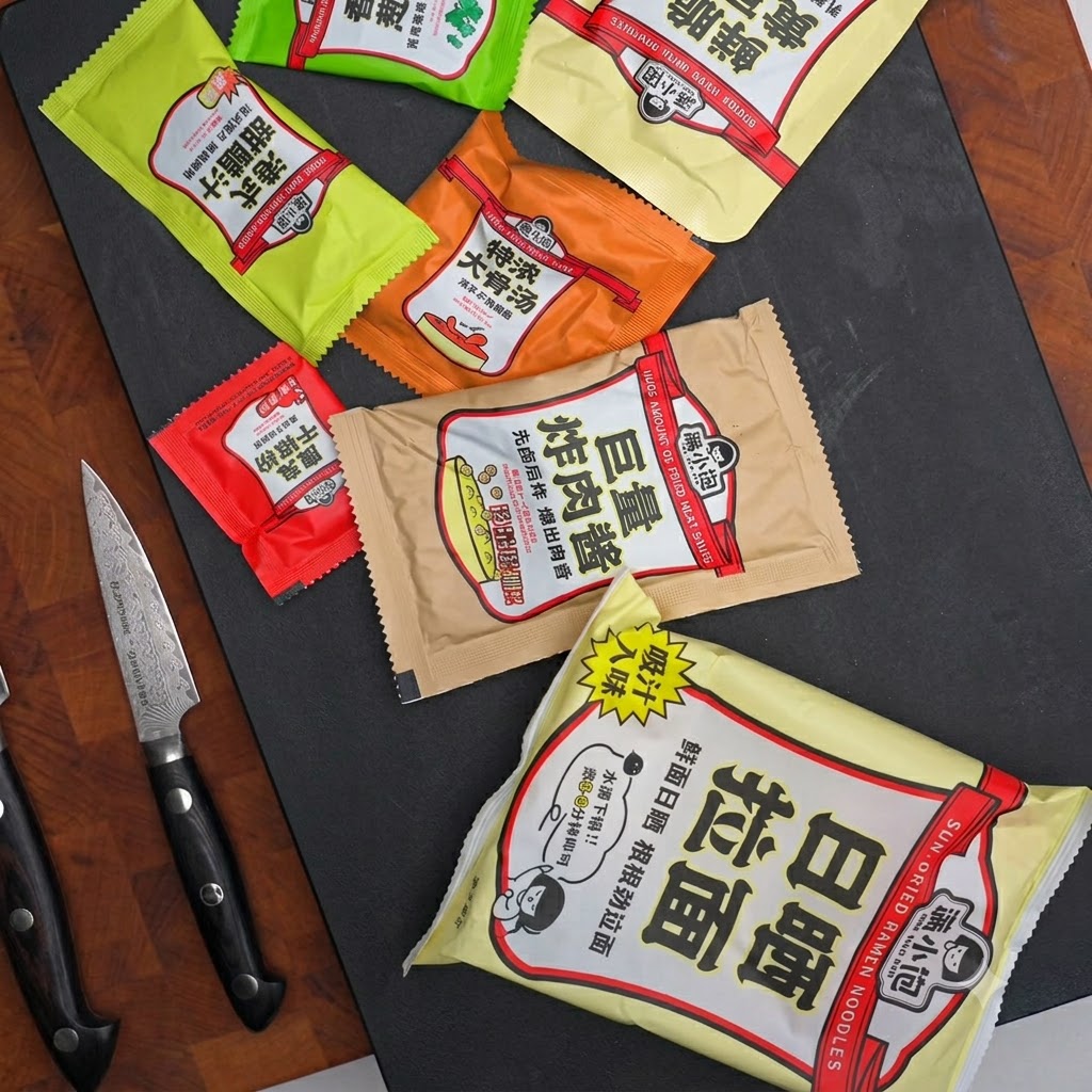Divers sachets dassaisonnement, sachets de sauce et un couteau disposés sur une surface sombre.