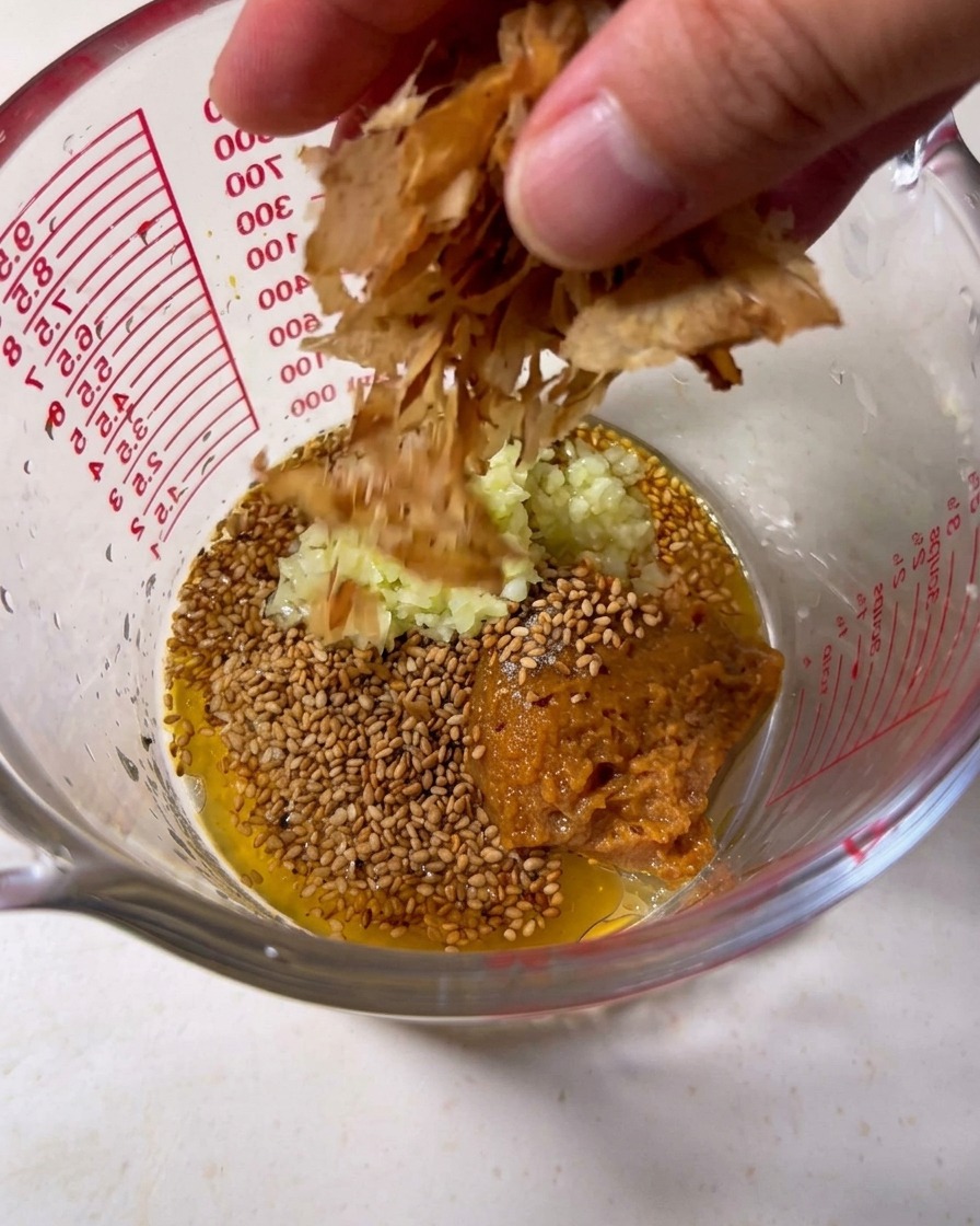 Le bouillon tonkotsu est versé dans un verre doseur rempli de graines de sésame, dail émincé et de flocons de bonite.