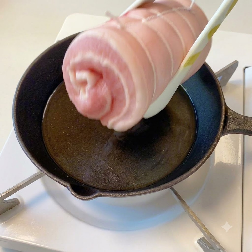 Le cylindre de poitrine de porc ficelé tenu par des pinces au-dessus dune poêle chaude contenant de lhuile.
