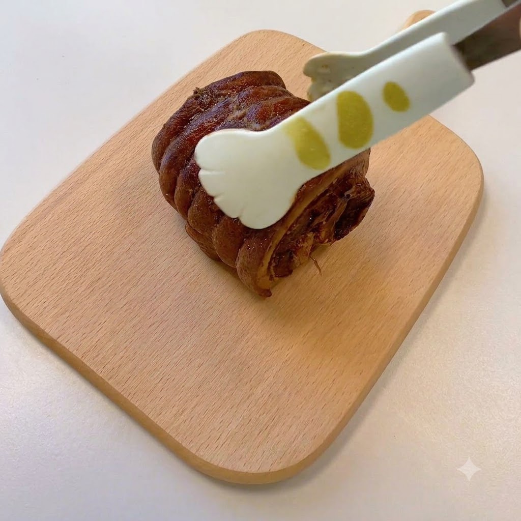 Une paire de pinces tenant un morceau de porc chashu cuit et roulé posé sur une planche à découper en bois.