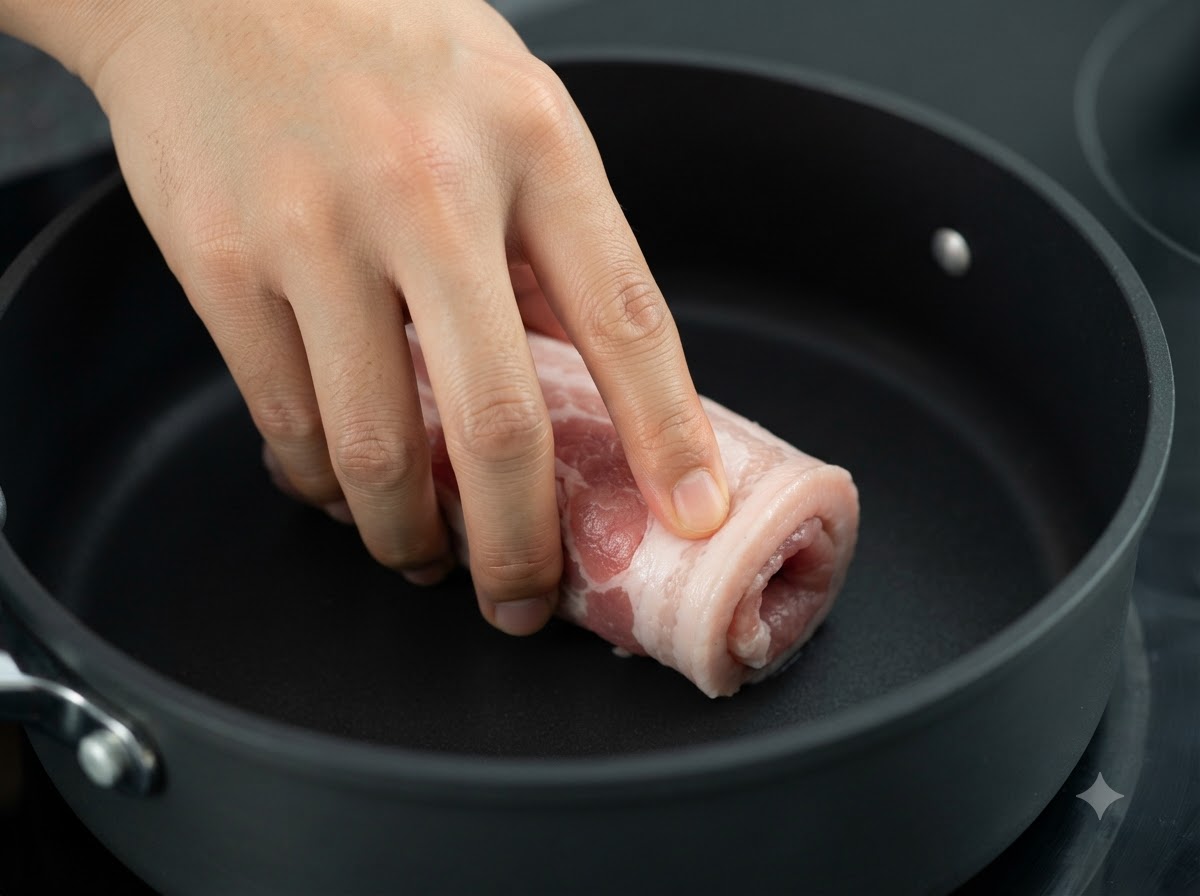 Le rouleau de poitrine de porc ficelé en train de dorer dans une poêle sombre sur le feu.